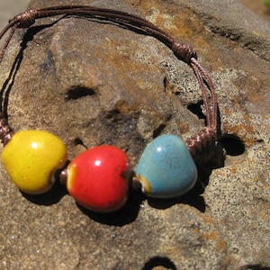May include: A handmade bracelet featuring three heart-shaped ceramic beads in yellow, red, and blue. The beads are strung on a brown cord with adjustable knots. The bracelet is displayed on a textured stone surface.