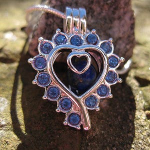 May include: A silver-colored heart-shaped pendant necklace with a small heart inside. The pendant is adorned with blue gemstones around the outer edge and holds a dark blue stone. The necklace is displayed on a textured, brown surface.