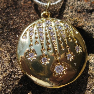 Puede incluir: Un colgante redondo de oro con un diseño celestial que presenta múltiples piedras claras y brillantes dispuestas en forma de estrella. El colgante cuelga de una cadena plateada y muestra una superficie metálica pulida y brillante.