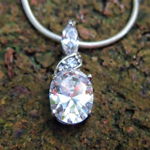 Pode incluir: Um colar de prata com um pingente brilhante. O pingente apresenta uma grande pedra preciosa clara de corte oval, acentuada com pedras claras menores. O colar é exibido em um fundo marrom texturizado.