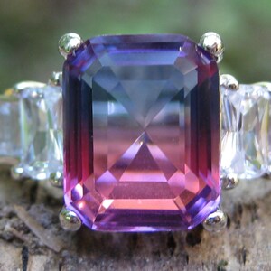 May include: A silver ring featuring a large, rectangular, color-shifting gemstone with shades of purple, pink, and blue. Flanking the central stone are smaller, clear, rectangular accent stones. The ring is set on a natural, textured surface.
