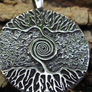 May include: A silver-colored pendant with a tree of life design. The pendant features a textured background and a detailed tree with roots, a trunk that spirals, and branches. The pendant is round and has a loop at the top for a chain.