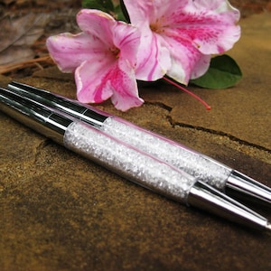 May include: Two silver pens with clear crystal accents. The pens are lying on a brown, textured surface.