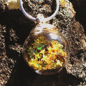 May include: A clear glass pendant necklace filled with gold star-shaped confetti. The pendant is suspended from a silver chain. The background is a dark, textured surface.