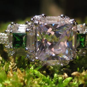Puede incluir: Un anillo plateado con una gran piedra preciosa cuadrada y transparente en el centro, flanqueada por dos piedras verdes cuadradas más pequeñas. El anillo está engastado con piedras transparentes más pequeñas y descansa sobre musgo verde.
