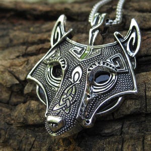 May include: A silver wolf head pendant necklace. The pendant has detailed patterns and dark accents for the eyes. The necklace is displayed on a textured, brown surface. The chain is a fine, silver link.