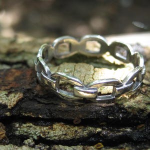 Könnte beinhalten: Ein silberfarbener Ring mit einem durchgehenden Kettenglied-Design. Das Band besteht aus einer Reihe von miteinander verbundenen, polierten Metallgliedern, die eine kreisförmige Form bilden und ein gleichmäßiges, sich wiederholendes Muster erzeugen.