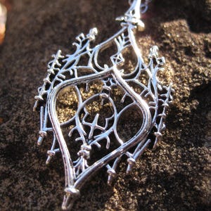 May include: Silver-toned pendant necklace with an intricate, openwork design. The pendant features a symmetrical, stylized floral or botanical pattern with delicate, branching details. The necklace is displayed against a textured, dark background.