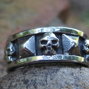 Może przedstawiać: Srebrny pierścień z motywem czaszki. Pierścień ma obręcz z szeregiem trójkątnych kształtów i czaszek.