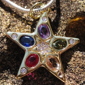 Peut inclure: Un pendentif en forme d'étoile dorée orné de pierres précieuses colorées, notamment bleues, violettes, rouges et vertes, serties dans un cadre doré. Le pendentif est attaché à une chaîne argentée, présenté sur une surface texturée.