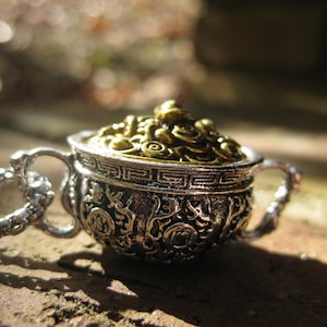 May include: A silver and gold charm necklace featuring a detailed ornate pot with a lid. The pot is filled with gold-colored decorative elements.