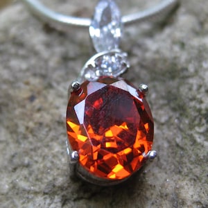 May include: A silver pendant necklace featuring a large, faceted, oval-cut, red-orange gemstone. The gemstone is set in a silver bezel and has a brilliant, reflective surface. The pendant is suspended from a silver chain, showcasing a classic jewelry design.