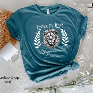 A teal t-shirt with a lion graphic and text, along with a mug, a woven basket, and some flowers, all arranged on a white surface.