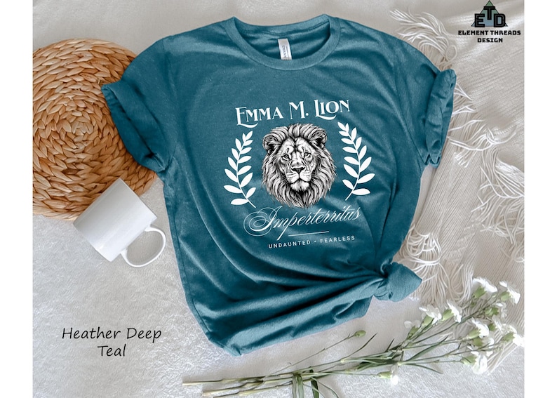 A teal t-shirt with a lion graphic and text, along with a mug, a woven basket, and some flowers, all arranged on a white surface.
