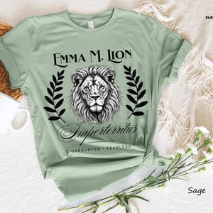 A green t-shirt with a lion graphic and text, along with a mug and some flowers, is displayed on a white surface.