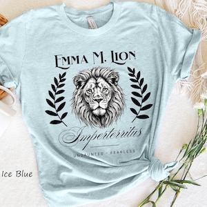 A light blue t-shirt with a graphic design featuring a lion's head and laurel leaves, along with the text "Emma M. Lion" and "Unbounded - Fearless". The t-shirt is placed on a white surface, and there is a woven basket and a bouquet of white flowers nearby.