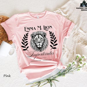 A pink t-shirt with a lion graphic and text, along with a mug and some flowers, is displayed on a white surface.