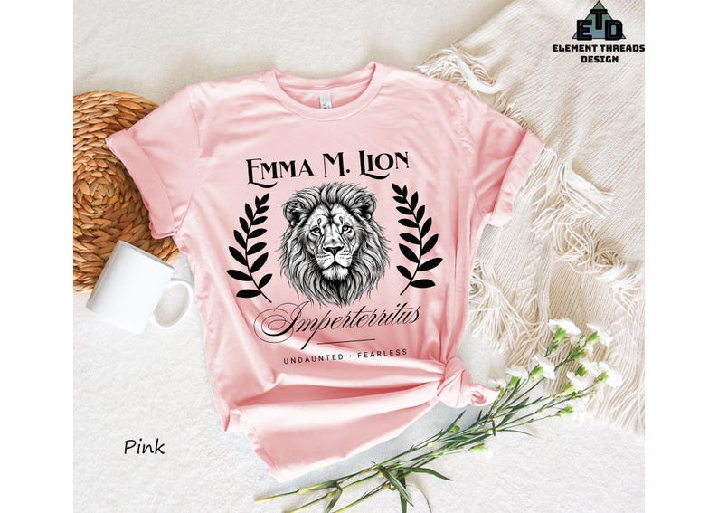 A pink t-shirt with a lion graphic and text, along with a mug and some flowers, is displayed on a white surface.