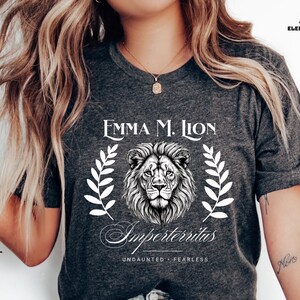 A woman wearing a gray t-shirt with a lion graphic and text, posing with her hair blowing in the wind.