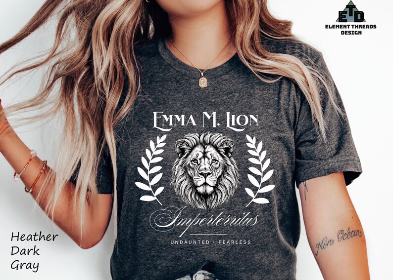 A woman wearing a gray t-shirt with a lion graphic and text, posing with her hair blowing in the wind.