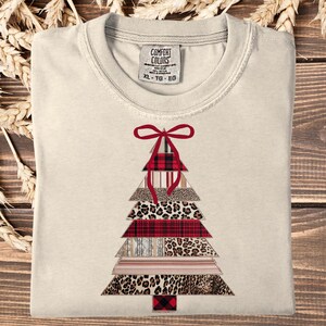 May include: Ivory t-shirt with a Christmas tree design. The tree is made of layered triangles with patterns like leopard print and plaid. A red bow adorns the top. The shirt has a crew neck and short sleeves. The label reads 'Comfort Colors'.