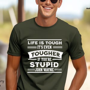 May include: Military green t-shirt with the quote "LIFE IS TOUGH IT'S EVEN TOUGHER IF YOU'RE STUPID JOHN WAYNE" in white block letters. The shirt has a crew neck and short sleeves. The Element Threads Design logo is in the upper right corner.