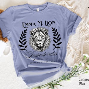 A light blue t-shirt with a graphic design featuring a lion's head and the text "Emma M. Lion" and "Emperortherius" printed on it. The t-shirt is placed on a white surface, with a white mug and a white woven basket nearby.