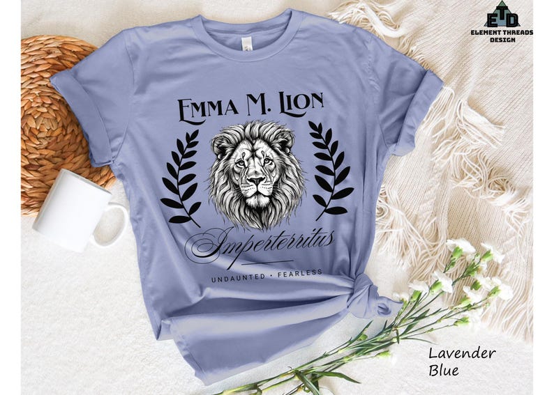 A light blue t-shirt with a graphic design featuring a lion's head and the text "Emma M. Lion" and "Emperortherius" printed on it. The t-shirt is placed on a white surface, with a white mug and a white woven basket nearby.