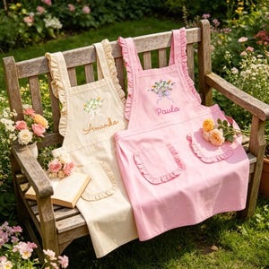 May include: Two aprons, one cream and one pink, with ruffled edges and pockets, rest on a wooden bench. The cream apron has "Amanda" embroidered, and the pink one says "Paula." Both feature floral embroidery. Roses and a book are nearby.