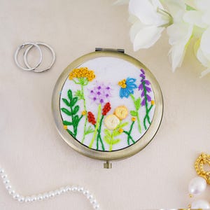 May include: A round, bronze-toned compact mirror featuring a white fabric insert embroidered with a colorful wildflower meadow design. The embroidery includes yellow, blue, purple, red, and cream-colored floral motifs with green stems.