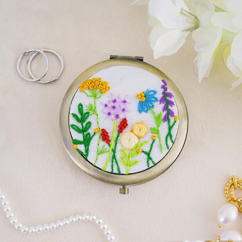 May include: A round, bronze-toned compact mirror featuring a white fabric insert embroidered with a colorful wildflower meadow design. The embroidery includes yellow, blue, purple, red, and cream-colored floral motifs with green stems.