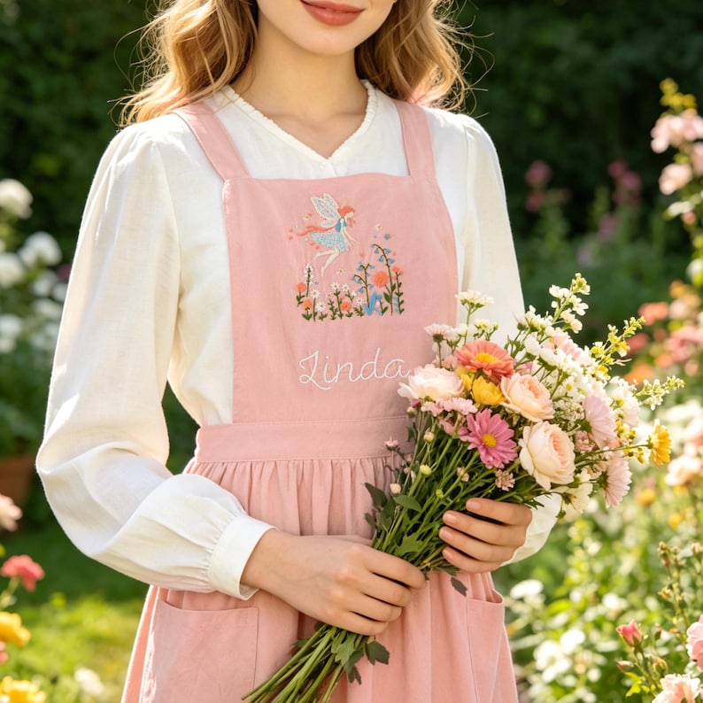 May include: A dusty pink pinafore apron featuring an embroidered fairy motif above a garden scene and the name Linda stitched in white cursive script. The apron has wide shoulder straps and a gathered skirt with visible pockets at the waist.