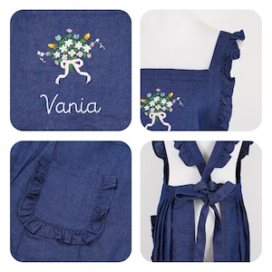 May include: A denim apron with a floral embroidery design and the name "Vania" in script. The apron features a ruffled pocket and straps, and a bow in the back. The embroidery includes white, yellow, blue, and pink flowers.