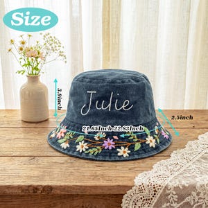 May include: A dark blue denim bucket hat featuring the embroidered name Julie in white script and a colorful floral pattern along the brim. The hat measures 3.9 inches in height, 2.5 inches for the brim, and 21.65 to 22.83 inches in circumference.
