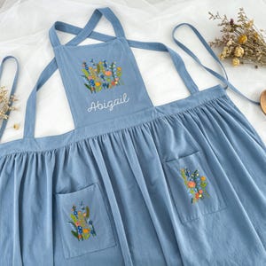 May include: A dusty blue apron featuring a gathered skirt and cross-back straps. The chest panel displays a colorful embroidered wildflower bouquet above the name Abigail in white script. Two front pockets each feature matching floral embroidery.