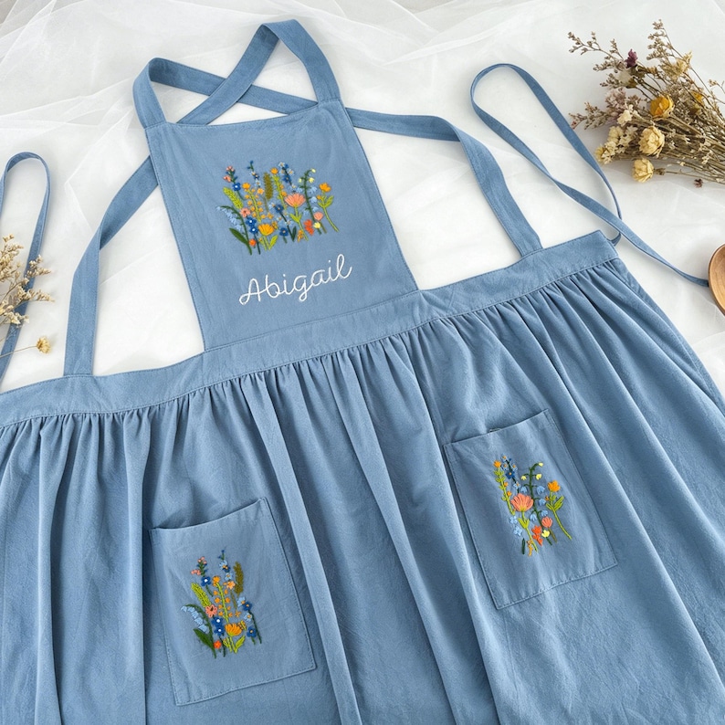 May include: A dusty blue apron featuring a gathered skirt and cross-back straps. The chest panel displays a colorful embroidered wildflower bouquet above the name Abigail in white script. Two front pockets each feature matching floral embroidery.