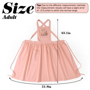 May include: A pink adult apron featuring a floral and fairy graphic with the name "Lynda" embroidered on the chest. The apron has cross-back straps and two front pockets. Dimensions are 109.47 cm in length and 131.83 cm in width.