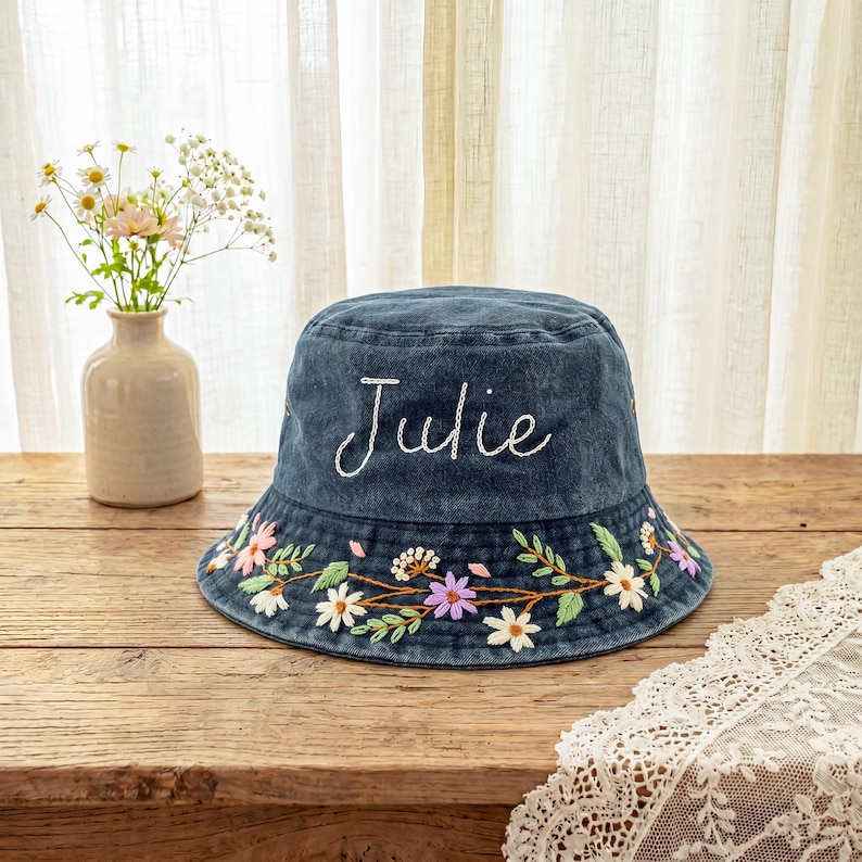 May include: A dark blue denim bucket hat featuring the name Julie embroidered in white script on the front. The brim is decorated with an embroidered floral vine pattern in shades of white, pink, purple, and green with brown stems.