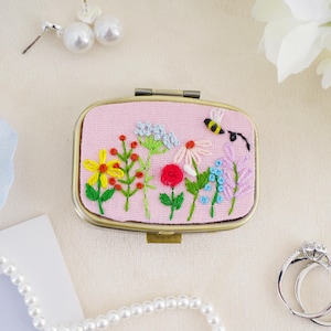May include: A rectangular metal compact case with a pink fabric top featuring hand-embroidered colorful wildflowers and a small bee. The embroidery includes yellow, red, blue, and purple floral stems with green leaves on a light pink background.