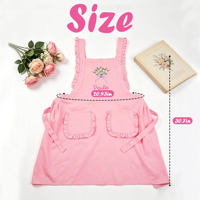 May include: A pink apron with ruffled edges and two front pockets. The apron has a floral design and the name "Paula" printed on it. The apron is 20.93 inches wide and 30.7 inches long. The word "Size" is written in pink.