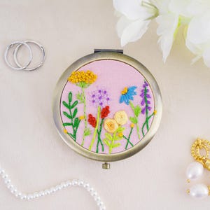 May include: A round metal compact mirror featuring a light pink fabric cover with hand-embroidered wildflowers in yellow, blue, purple, and red. The floral design includes green stems and leaves, with a metallic frame and a hinge at the top.