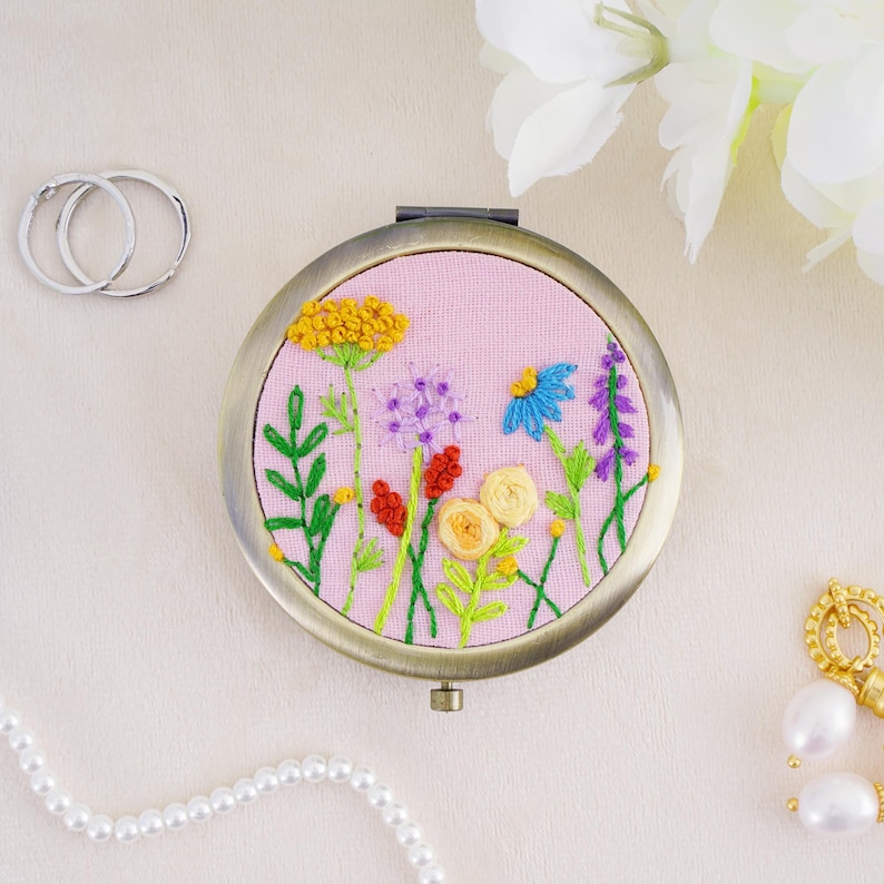 May include: A round metal compact mirror featuring a light pink fabric cover with hand-embroidered wildflowers in yellow, blue, purple, and red. The floral design includes green stems and leaves, with a metallic frame and a hinge at the top.