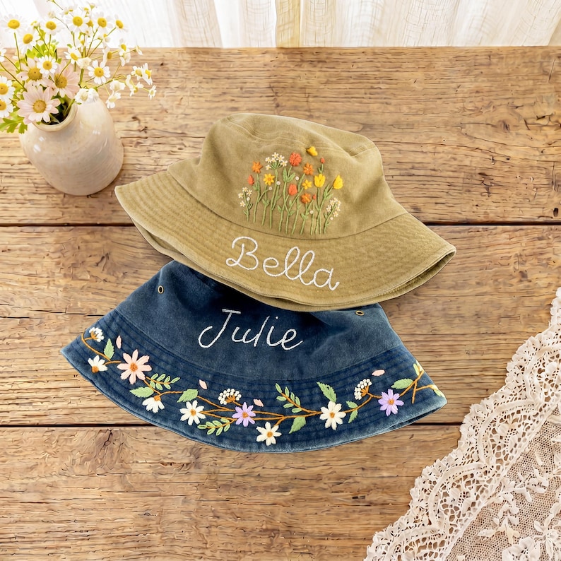 May include: An olive green bucket hat with embroidered wildflowers and the name Bella, stacked on a dark blue denim bucket hat featuring an embroidered floral vine border and the name Julie in white script lettering.