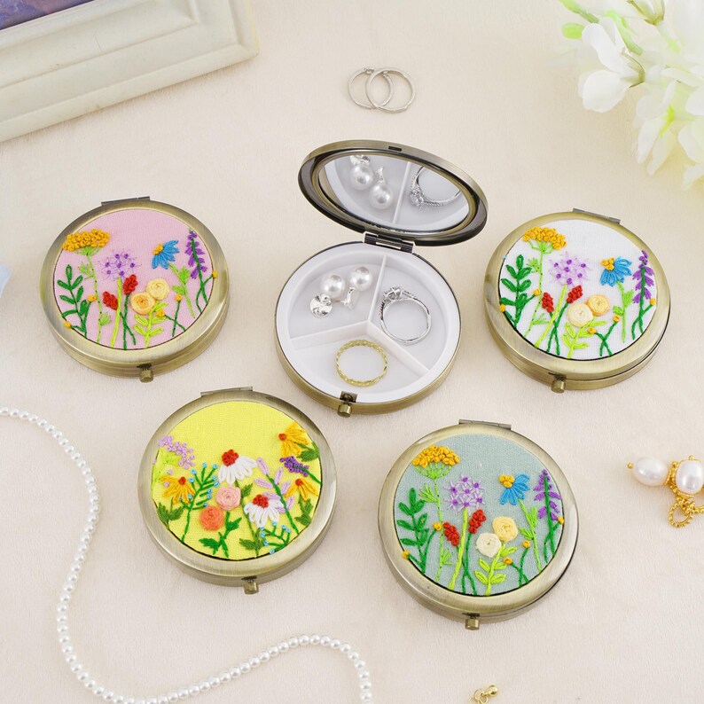 May include: Four round metal jewelry cases with hinged lids featuring embroidered floral designs in pink, white, yellow, and light blue. One case is open, revealing a white interior divided into three sections containing silver rings and pearl earrings.