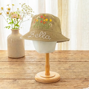 May include: A tan bucket hat featuring embroidered colorful wildflowers on the crown and the name Bella stitched in white cursive script on the brim. The hat is displayed on a stand, showcasing the detailed floral embroidery and white lettering.