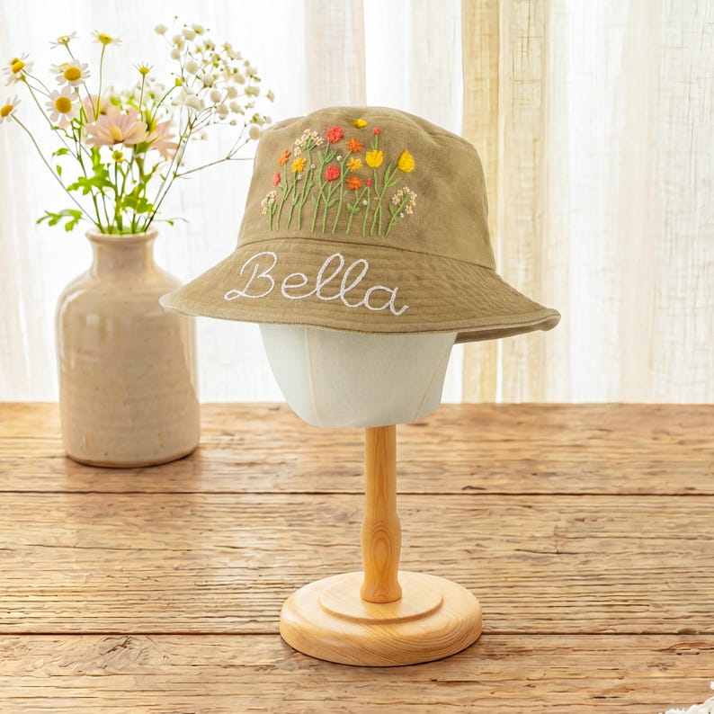 May include: A tan bucket hat featuring embroidered colorful wildflowers on the crown and the name Bella stitched in white cursive script on the brim. The hat is displayed on a stand, showcasing the detailed floral embroidery and white lettering.