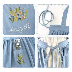 May include: A light blue apron featuring colorful floral embroidery and the name Abigail stitched in white thread. The apron includes a pocket with matching floral embroidery, long fabric waist ties, and shoulder straps for a secure fit.