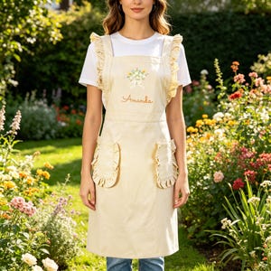 May include: A cream-colored apron with ruffled edges and straps. The apron features a floral embroidery and the name "Amanda" in cursive. Two ruffled pockets are on the front. The apron is worn over a white t-shirt.