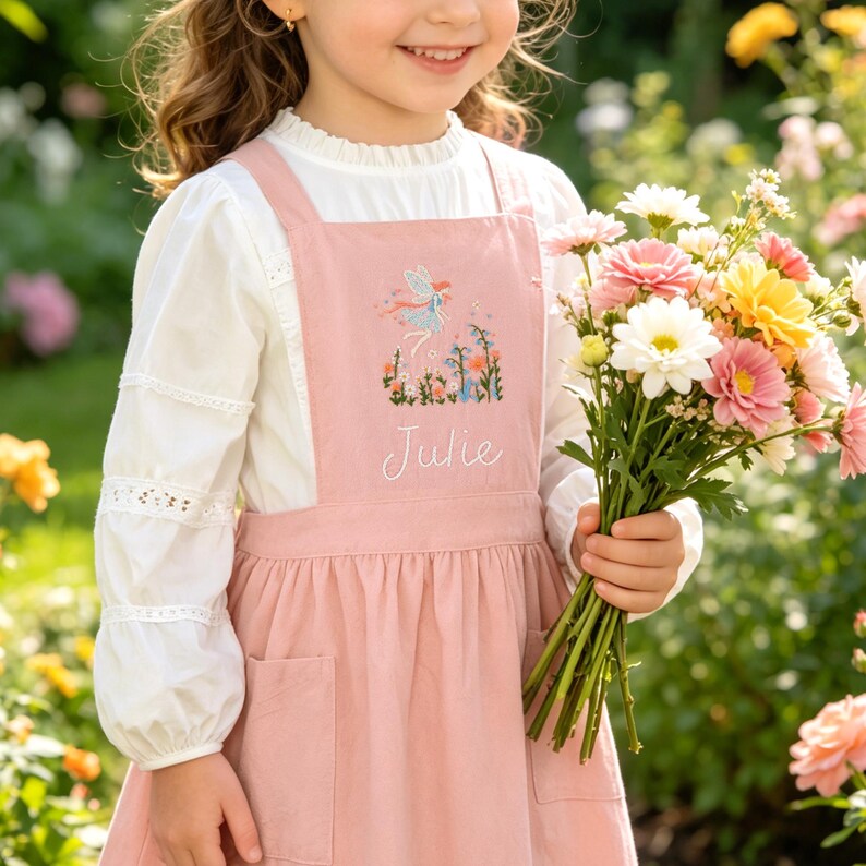 May include: A dusty pink pinafore dress featuring a white long-sleeved blouse with lace trim. The dress has a central embroidered design of a fairy and flowers above the white stitched name Julie, and includes two front patch pockets.