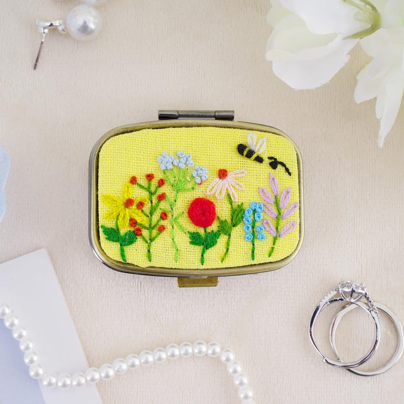 May include: An oval metal compact case featuring a yellow fabric top with hand-embroidered colorful wildflowers and a small black and white bee. The embroidery includes red, blue, green, and purple floral stems and blossoms on a bright yellow background.